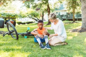 Woman kneeling beside a boy sitting on the grass next to his fallen bicycle after a bicycle accident
