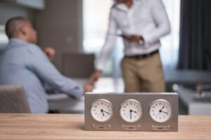 World-clock display showing Dubai, Sofia, and London times on an office desk with two people in the background, symbolizing how long an auto accident settlement takes to resolve